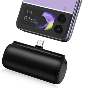 A compact wireless charger with a USB-C connector is positioned near a smartphone with a small external display showing the time and date, and dual camera lenses.