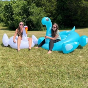 Two inflatable pool toys, one shaped like a swan and the other like a dinosaur, with two people sitting beside them.