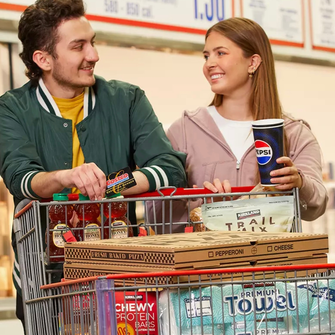 A man and woman smile over a shopping cart filled with Costco items, including honey, fresh baked pizza, trail mix, chewy protein bars, Pepsi, and premium towels. The man holds a Costco membership card.