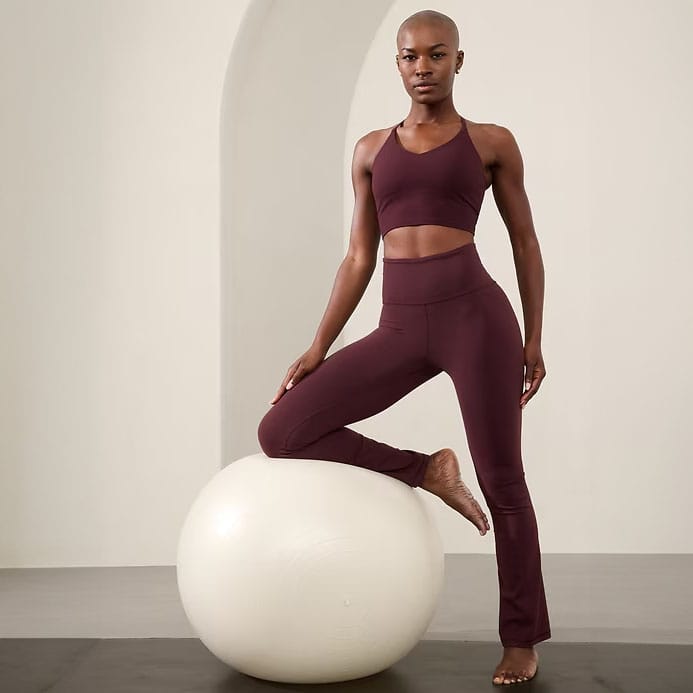 A person is wearing a matching maroon activewear set consisting of high-waisted leggings and a sports bra, posing with one foot on a white exercise ball.