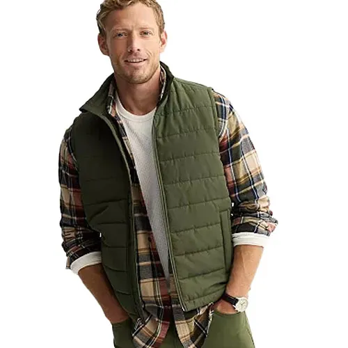 A man is wearing an olive green quilted vest over a plaid shirt with rolled-up sleeves, paired with a white undershirt. He is also wearing a wristwatch.