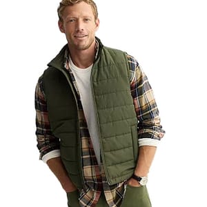 A man is wearing an olive green quilted vest over a plaid shirt with rolled-up sleeves, paired with a white undershirt. He is also wearing a wristwatch.