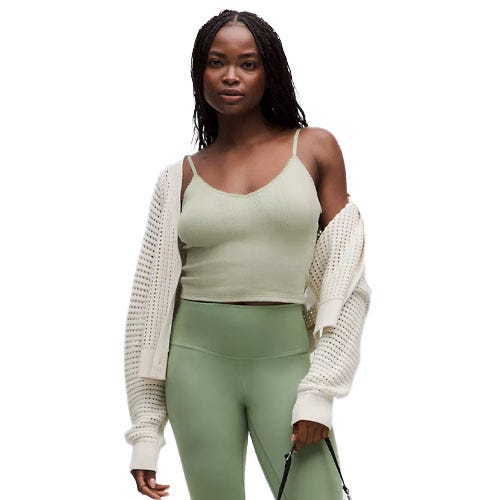 A woman is wearing a light green tank top, sage green high-waisted leggings, and a cream perforated cardigan draped over her shoulders.