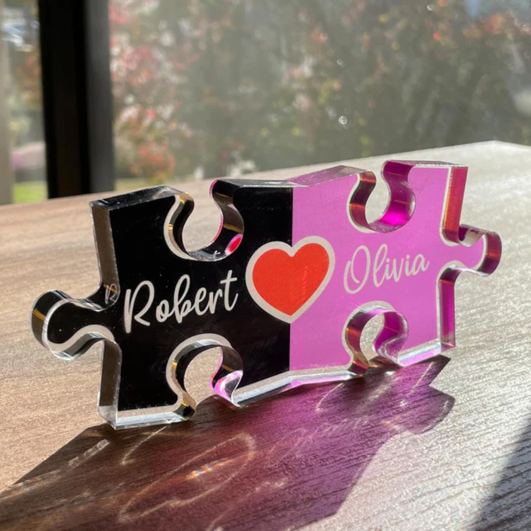 A puzzle piece-shaped acrylic block features the names \“Robert\“ and \“Olivia,\“ with a red heart symbol between them, divided into two colors: black and pink.