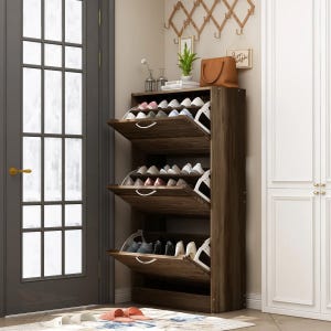 Wooden shoe rack with three drop-down compartments, holding multiple pairs of shoes. It is positioned near a door, with a decorative wall hanger and a small plant on top. A rug and additional footwear are on the floor.