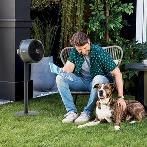 A man seated outdoors next to a standing fan and a dog.