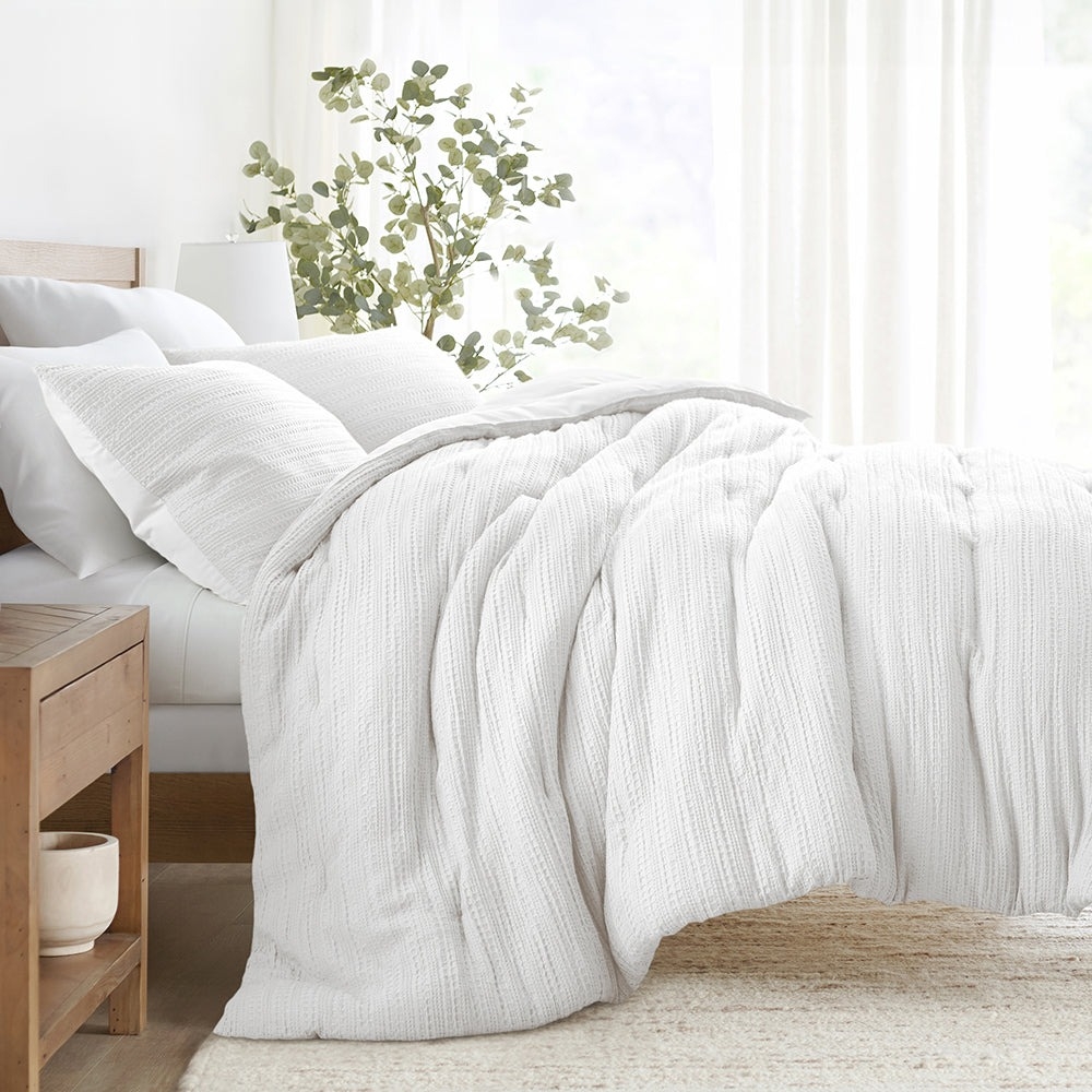 A cozy white bedding set featuring a textured comforter and matching pillow shams on a neatly arranged bed, with a wooden nightstand and decorative plant in the background.
