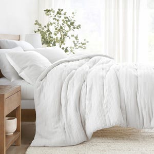 A cozy white bedding set featuring a textured comforter and matching pillow shams on a neatly arranged bed, with a wooden nightstand and decorative plant in the background.