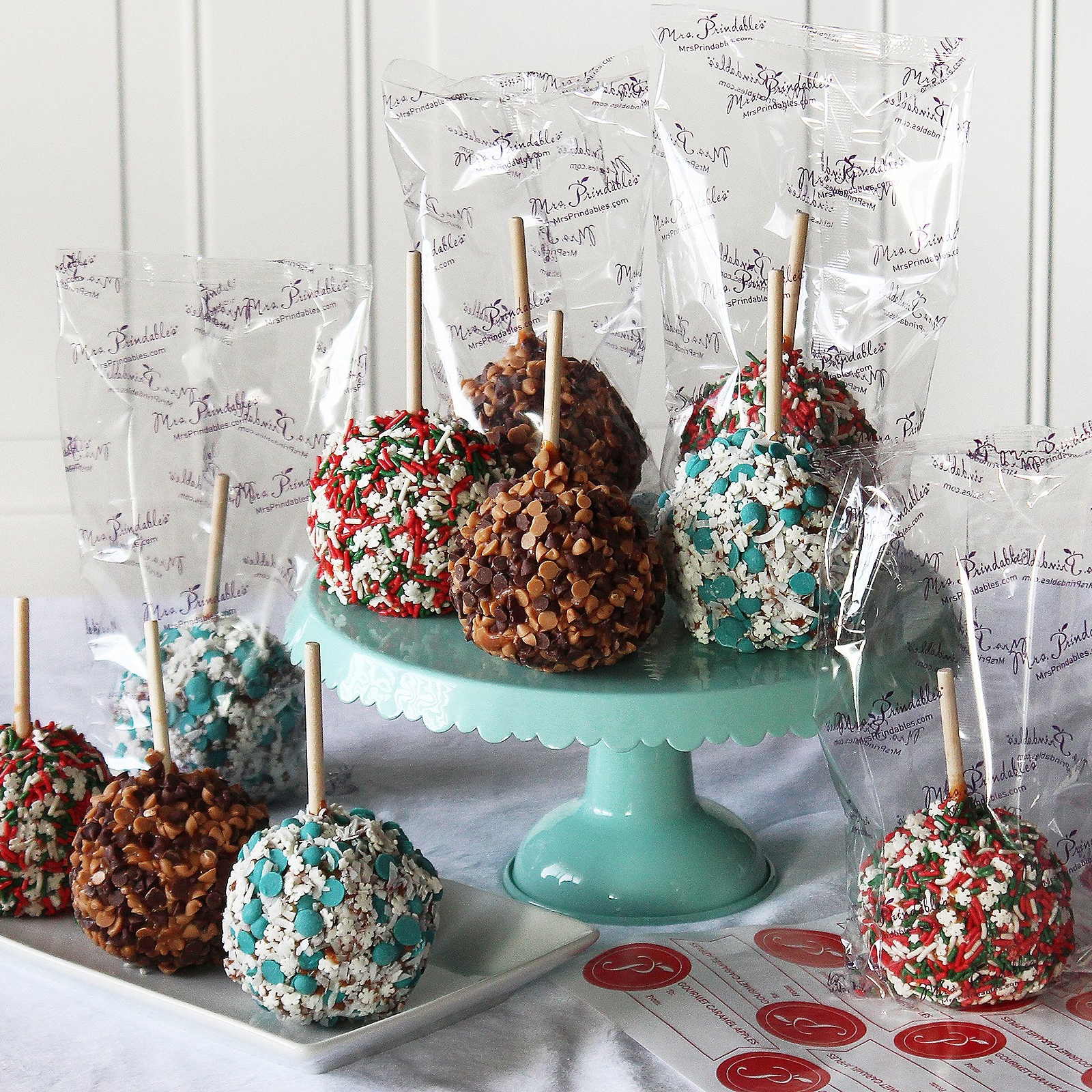 A set of 10 Mrs. Prindables caramel apples decorated with various toppings like chocolate chips and colorful sprinkles, individually wrapped in clear packaging with branding and arranged on a turquoise cake stand and white plate.