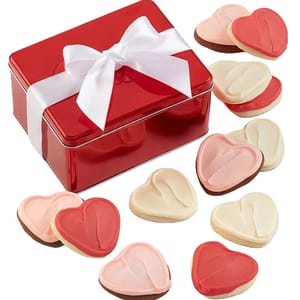 A red tin gift box with a white ribbon contains heart-shaped cookies, some frosted in red, white, and pink.