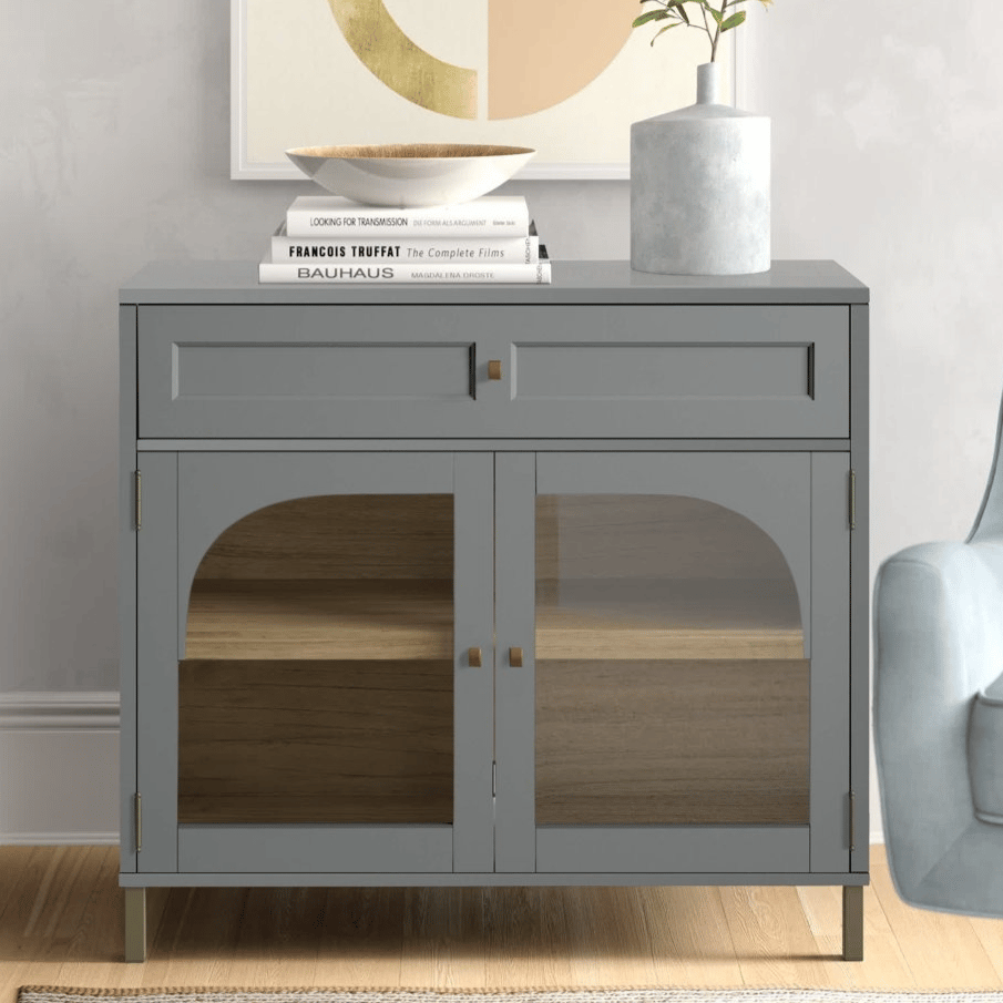 A gray cabinet with a glass front and brass details sits next to an armchair. The top is adorned with a stack of books, a bowl, and a vase with greenery.