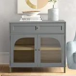 A gray cabinet with a glass front and brass details sits next to an armchair. The top is adorned with a stack of books, a bowl, and a vase with greenery.
