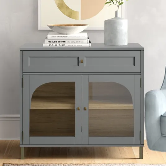 A gray cabinet with a glass front and brass details sits next to an armchair. The top is adorned with a stack of books, a bowl, and a vase with greenery.