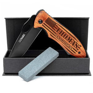 Folding knife with a wooden handle featuring engraved text and a design of the U.S. flag, accompanied by a sharpening stone, set in a black box.