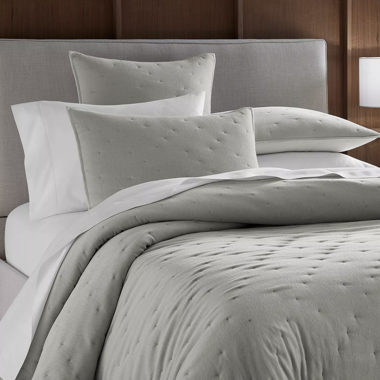 A neatly made bed with a light gray dotted duvet cover, matching pillows, and white sheets, set against a gray headboard and wooden wall backdrop.