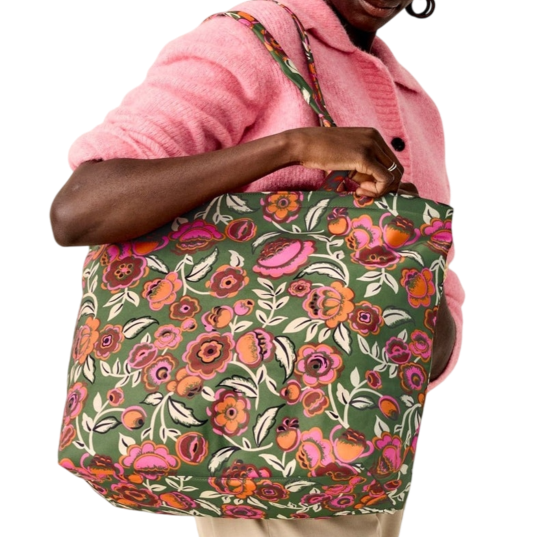A person wearing a pink sweater and beige pants holds a large tote bag with a colorful floral pattern on a green background.