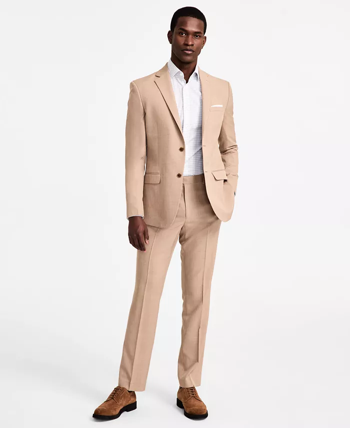 A man is dressed in a stylish tan suit with a single-breasted jacket and matching trousers, paired with a white shirt and brown suede shoes.