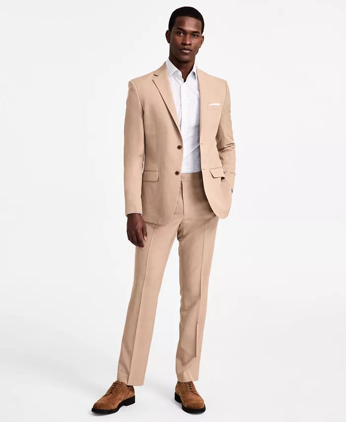 A man is dressed in a stylish tan suit with a single-breasted jacket and matching trousers, paired with a white shirt and brown suede shoes.