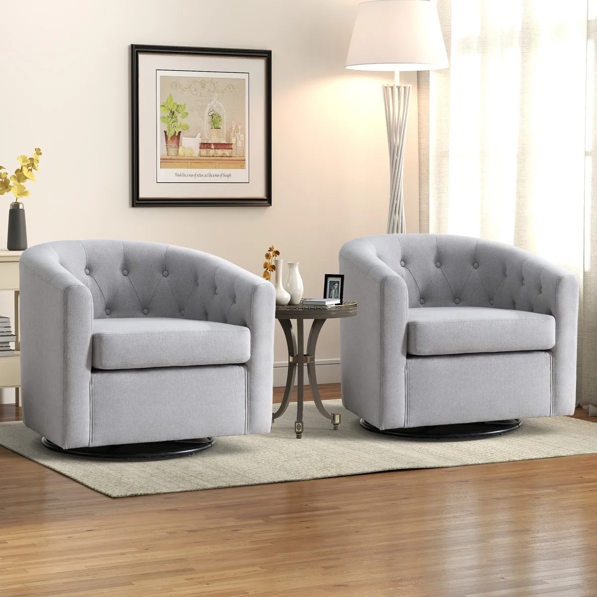 Two gray swivel armchairs with tufted backs are placed on a light rug, separated by a round side table with decorative items. A floor lamp and framed wall art complete the cozy living room setup.