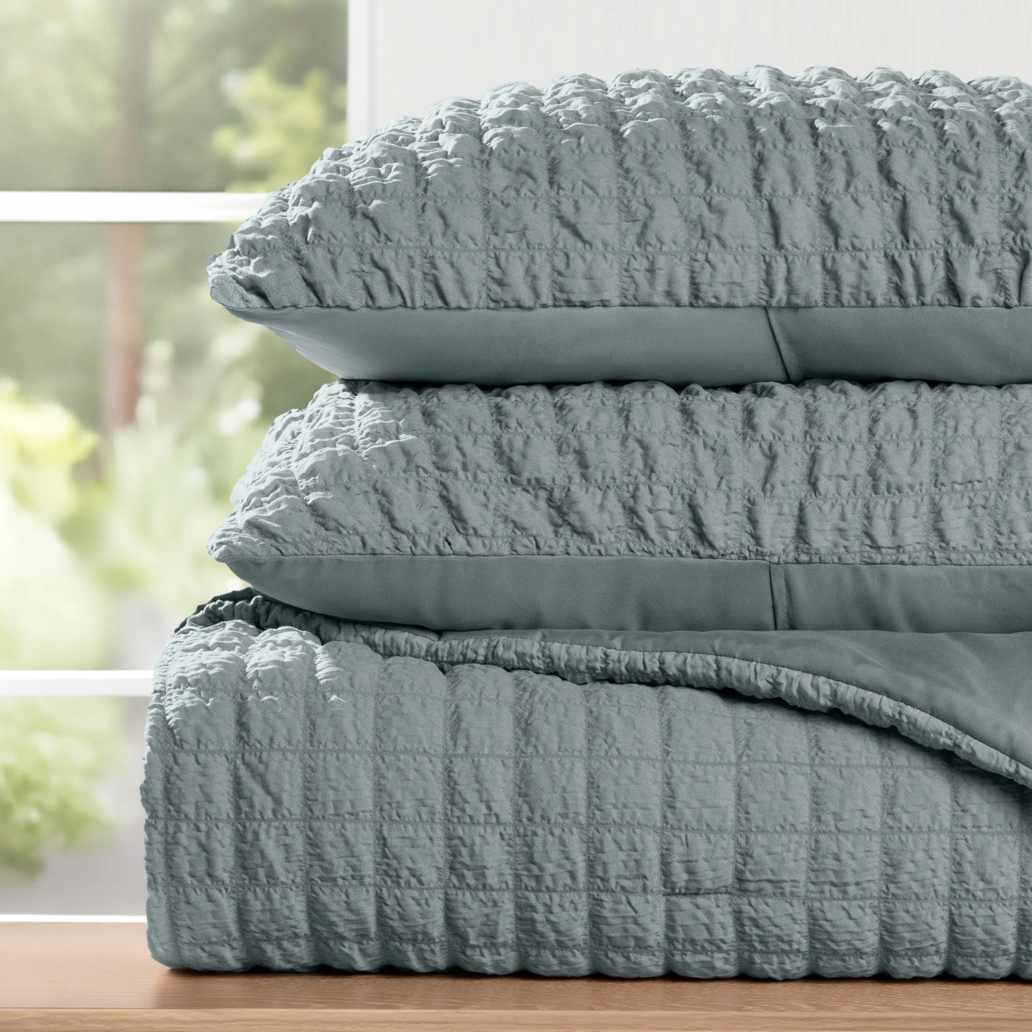A stack of textured, light gray bedding, including two pillows and a quilted comforter, is displayed on a wooden surface near a window.