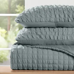 A stack of textured, light gray bedding, including two pillows and a quilted comforter, is displayed on a wooden surface near a window.