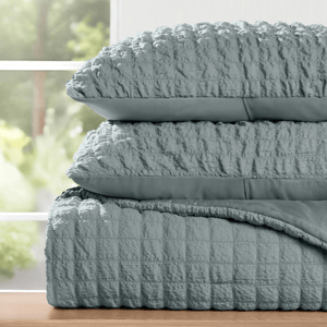 A stack of textured, light gray bedding, including two pillows and a quilted comforter, is displayed on a wooden surface near a window.