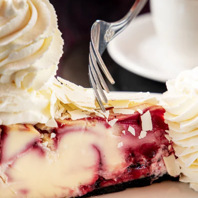A slice of cheesecake with swirls of cherry and a chocolate crust, topped with whipped cream and white chocolate shavings, is being cut with a fork. A cup is partially visible in the background.