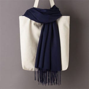 A white tote bag with handles is shown with a dark navy blue scarf draped over it. The scarf has a fringe at the bottom.