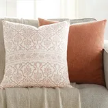A 20\“ pillow insert with a decorative case features a white background with a symmetrical, intricate floral pattern in light pink. It is paired with a muted orange pillow on a gray couch, adding a touch of elegance and warmth to the setting.