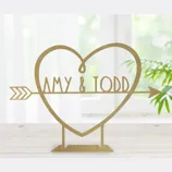 A decorative gold heart silhouette with the names \“AMY & TODD\“ intersected by an arrow, designed as a freestanding tabletop sign.