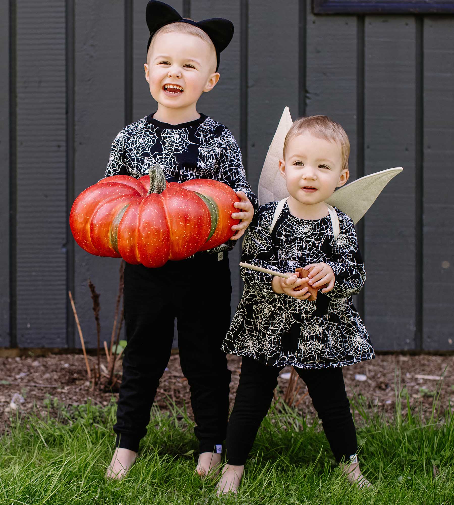 Two children dressed in matching spiderweb-patterned outfits; one wears cat ears and holds a large pumpkin, while the other has wings and holds a wand.