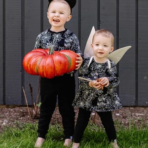Two children dressed in matching spiderweb-patterned outfits; one wears cat ears and holds a large pumpkin, while the other has wings and holds a wand.