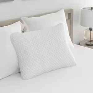White textured pillow on a bed with white bedding next to a bedside table with a lamp.