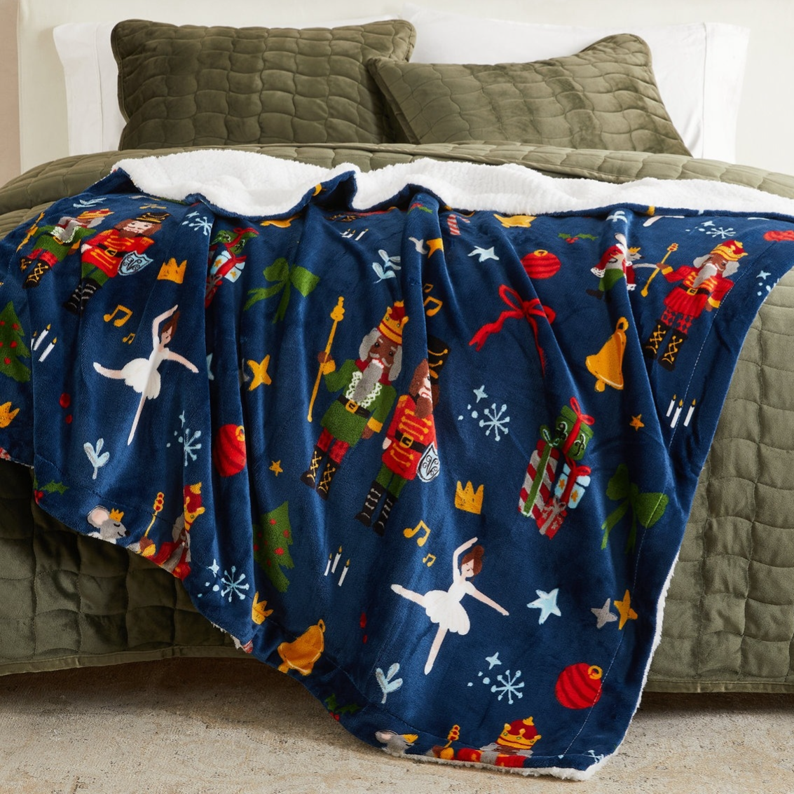 Fleece blanket with a Nutcracker theme, featuring soldiers, ballerinas, musical notes, and festive decorations, draped over an olive green quilted bedspread.