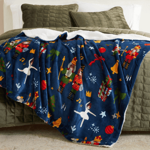 Fleece blanket with a Nutcracker theme, featuring soldiers, ballerinas, musical notes, and festive decorations, draped over an olive green quilted bedspread.