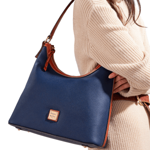 A navy Dooney & Bourke Pebble-Grain Leather Hobo with brown leather trim and a shoulder strap, featuring a label on the front. It's paired with a cream knit sweater.