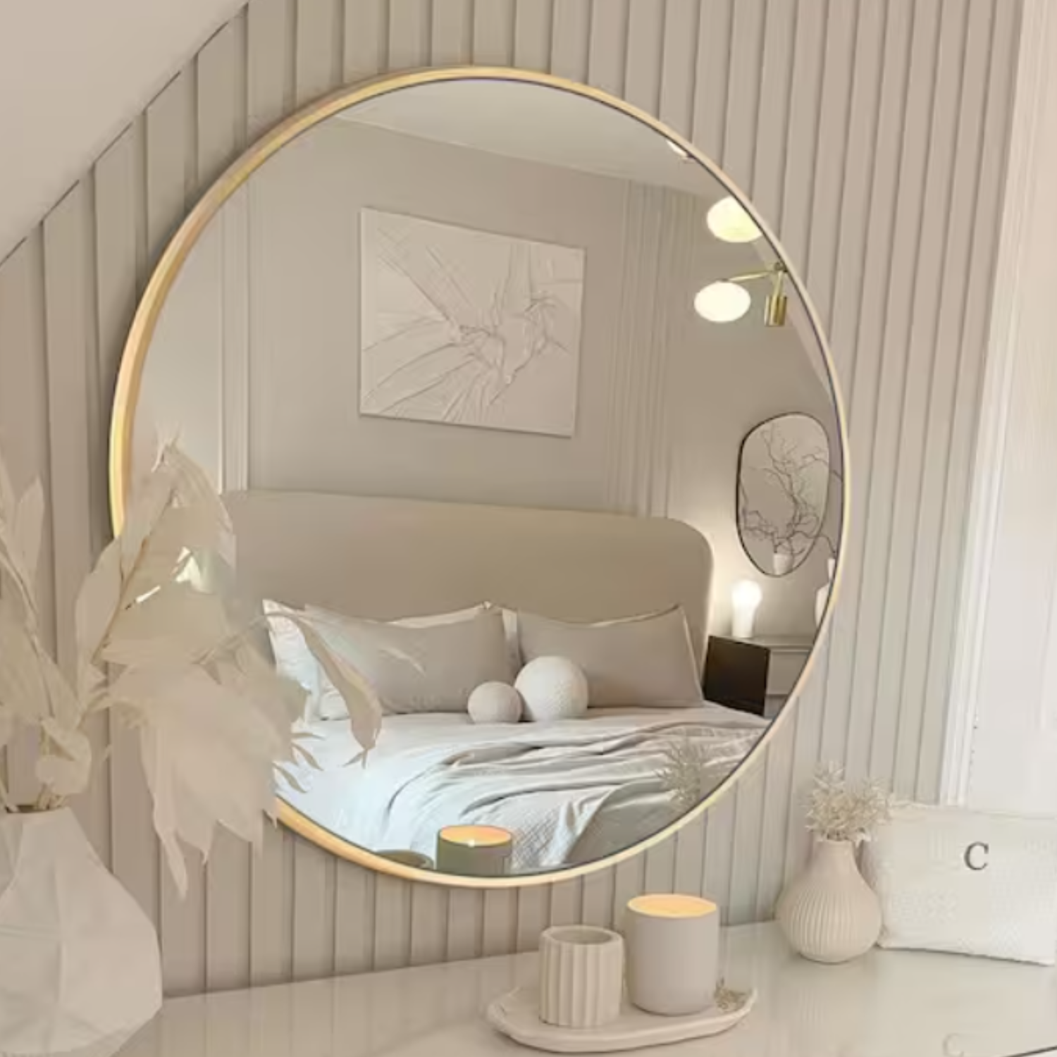 A round wall mirror with a slim gold rim is mounted on a vertically grooved wall, reflecting a minimalist bedroom setting with soft lightings, abstract art, and neutral bedding.