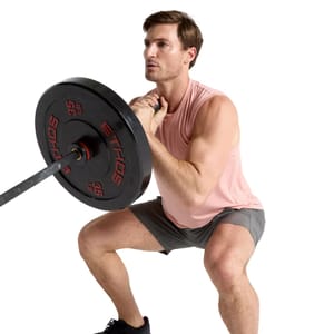 A man is performing a squat while wearing a sleeveless, light pink VRST Icon Tank and gray shorts. He is holding a barbell with black and red weights.