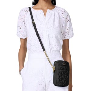 A person is wearing a white eyelet blouse and matching shorts, carrying a small black quilted crossbody bag with gold chain details.
