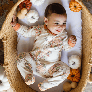 A baby in a woven basket wearing a floral-patterned sleeper with \“gobble gobble\“ text, surrounded by small decorative pumpkins.