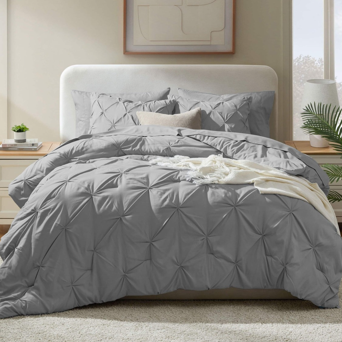 A neatly made bed features a grey, textured comforter set with matching pillow shams, a light brown accent pillow, and a white throw. The setting includes a bedside table with a small plant and books, in a room with soft lighting and a framed abstract print.