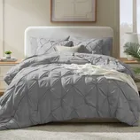 A neatly made bed features a grey, textured comforter set with matching pillow shams, a light brown accent pillow, and a white throw. The setting includes a bedside table with a small plant and books, in a room with soft lighting and a framed abstract print.