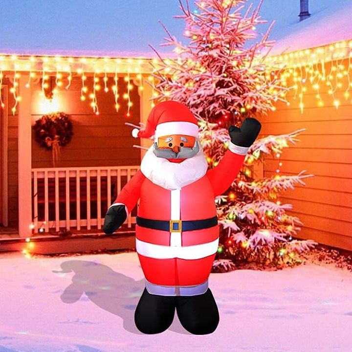 A six-foot-tall inflatable Santa Claus with a waving hand, wearing a red suit and a mask, is displayed outdoors with a snow-covered tree and holiday lights in the background.