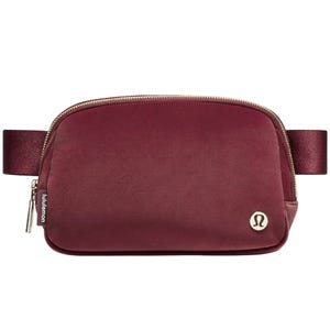 A maroon Lululemon crossbody bag with a zippered opening, featuring the Lululemon logo on the front and an adjustable strap.