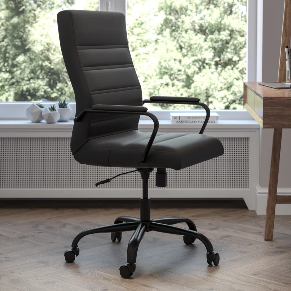 A black, high-back office chair with adjustable height and armrests sits on a five-wheel base next to a wooden desk with various stationery items. The room features a large window overlooking greenery.