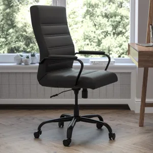 A black, high-back office chair with adjustable height and armrests sits on a five-wheel base next to a wooden desk with various stationery items. The room features a large window overlooking greenery.