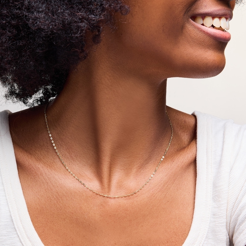 A close-up of a person wearing a delicate gold chain necklace. The individual has a wide smile and is dressed in a white top.