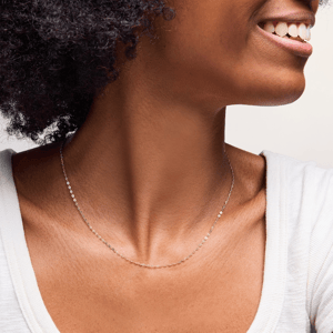 A close-up of a person wearing a delicate gold chain necklace. The individual has a wide smile and is dressed in a white top.