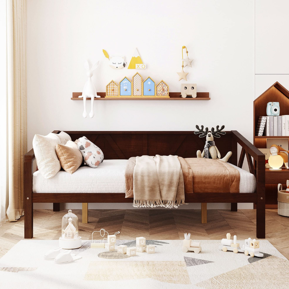 A cozy children's room features a wooden daybed with plush pillows and a soft blanket, a shelf with decorative items like toy houses and stuffed animals, and a rug with pastel-colored toys including a train set and building blocks.