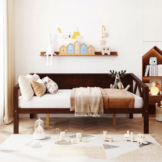 A cozy children's room features a wooden daybed with plush pillows and a soft blanket, a shelf with decorative items like toy houses and stuffed animals, and a rug with pastel-colored toys including a train set and building blocks.